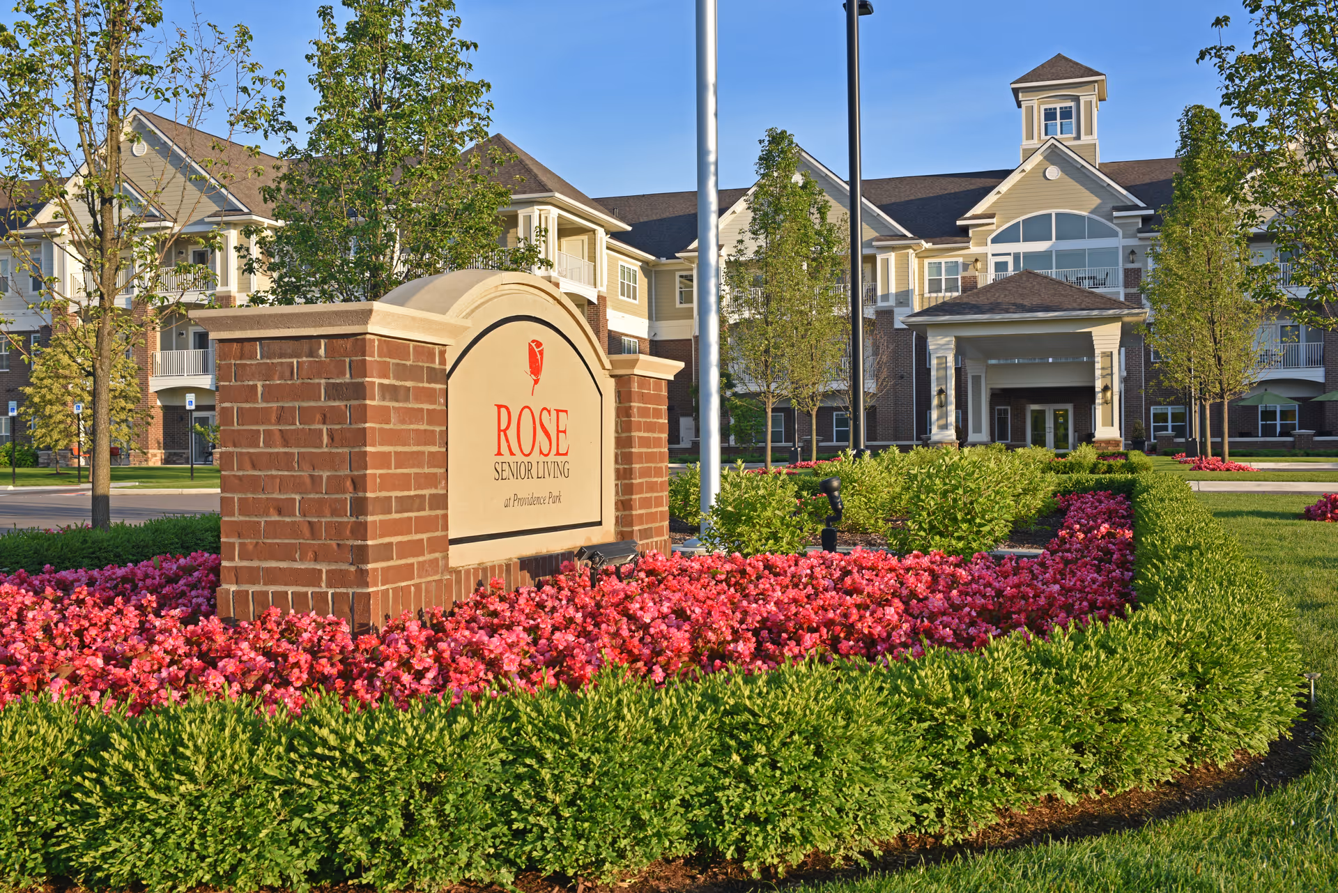 Exterior view of Rose Senior Living Providence Park facility with a brick sign surrounded by vibrant pink flowers and green shrubs in the foreground. The multi-story building with balconies and a covered entrance is visible in the background under a clear blue sky.
