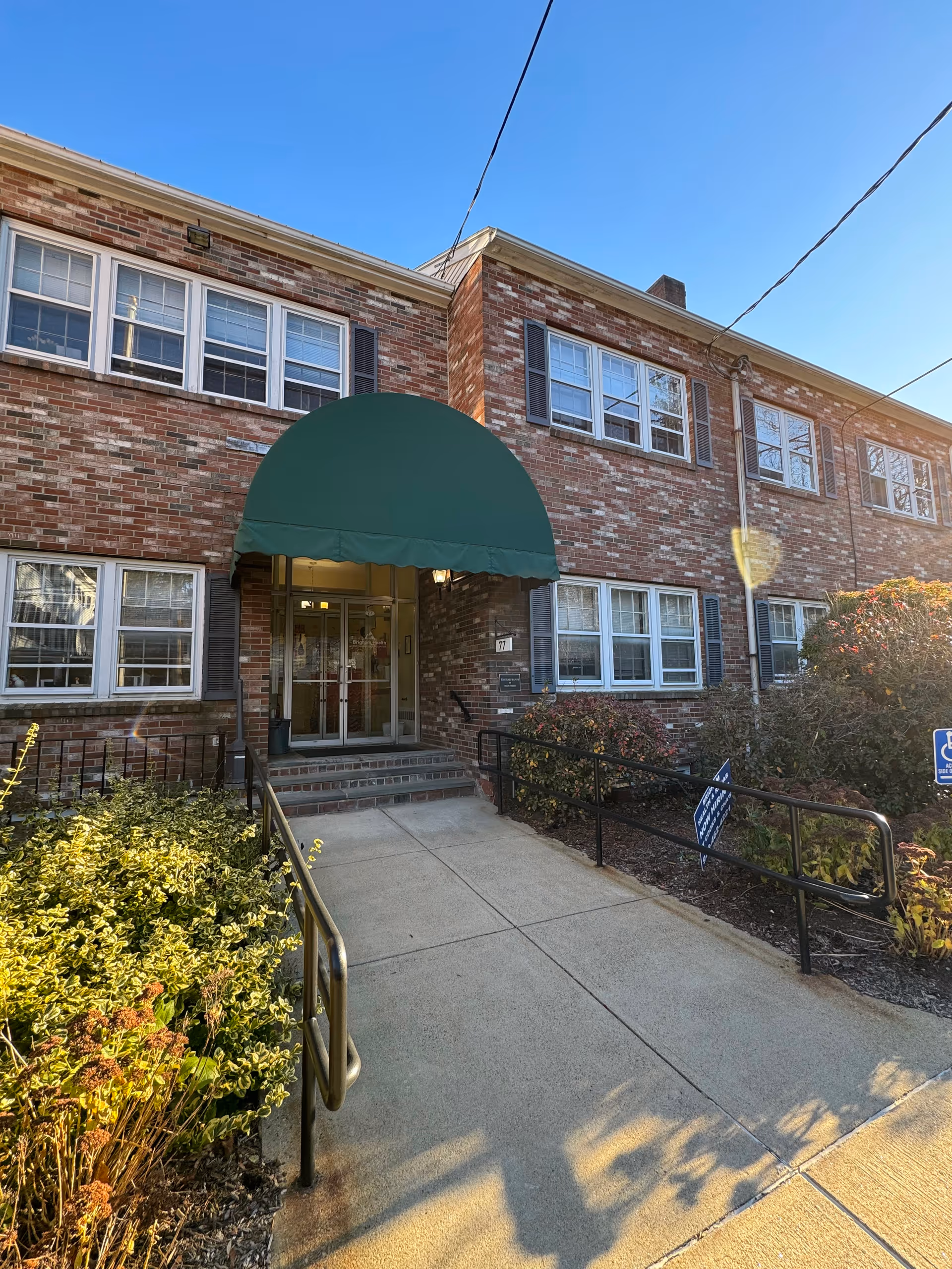 Two-story brick senior living facility exterior with green awning over entrance and ramp.