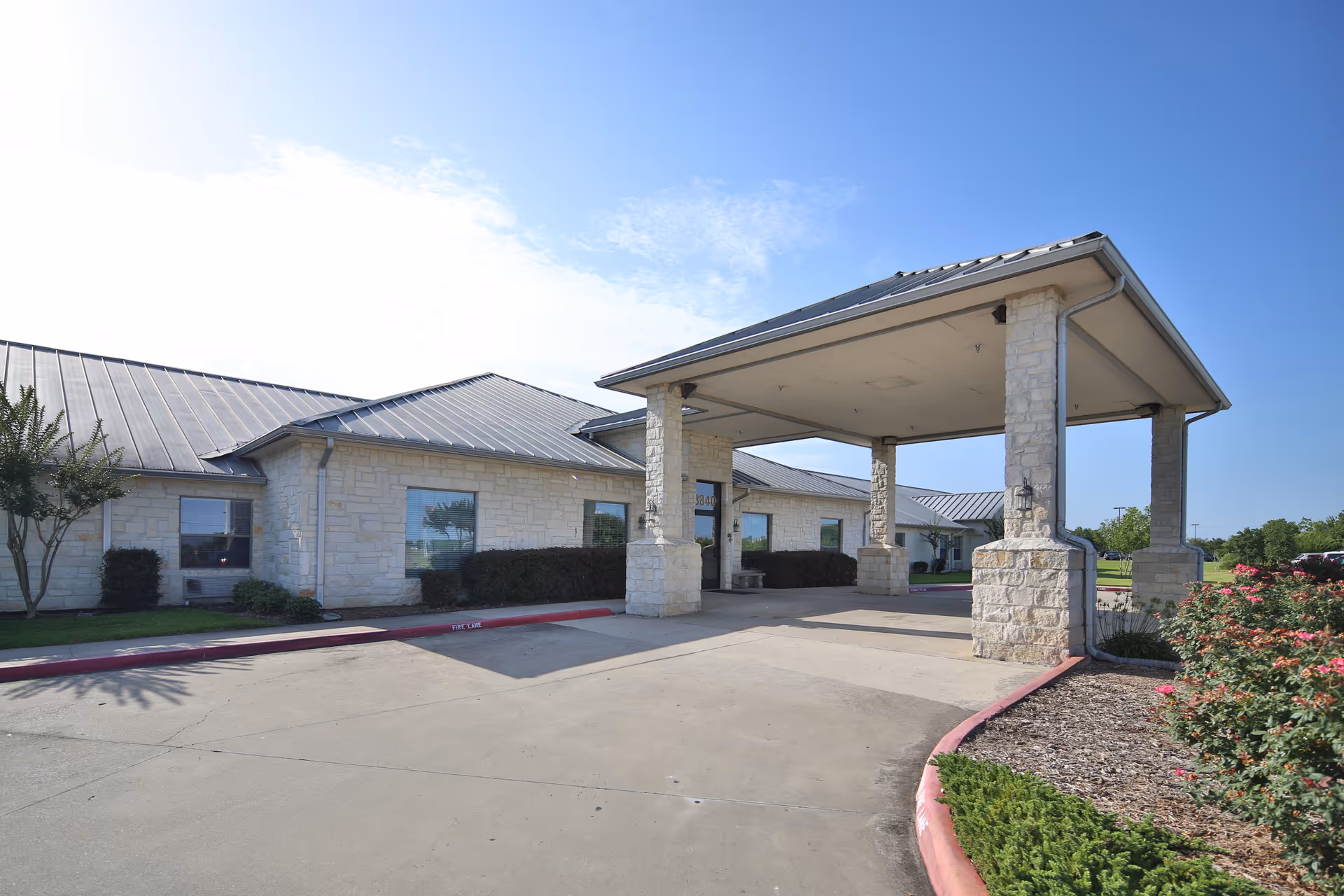 Exterior view of Jefferson Nursing and Rehabilitation Center showing a covered entrance with stone pillars, a driveway, and landscaped bushes and flowers under a clear blue sky.