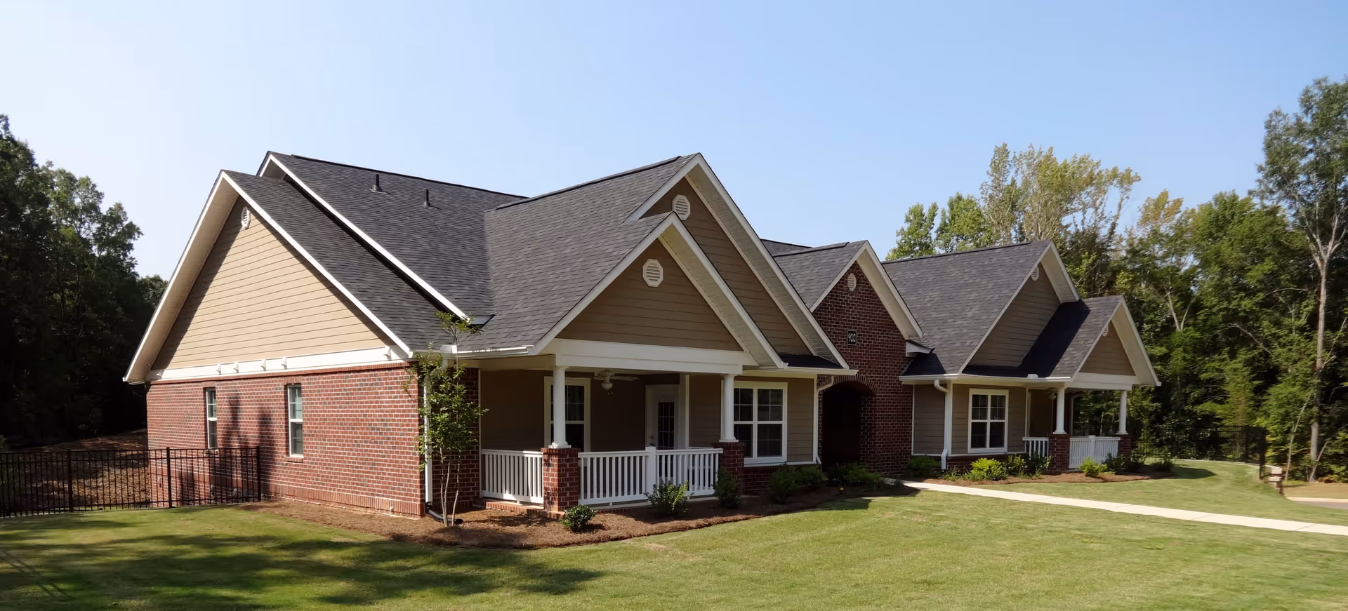 Exterior view of a single-story senior living facility building with a combination of brick and beige siding, multiple gabled roofs, white columns, and a small porch area. The building is surrounded by a well-maintained lawn and trees under a clear blue sky.