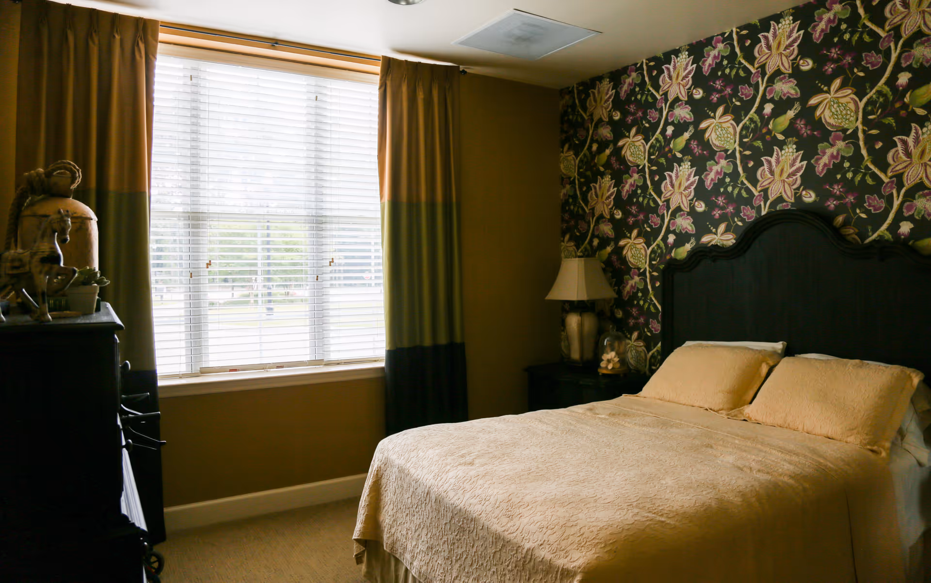 A cozy bedroom with a large window covered by blinds and curtains. The bed has a dark wooden headboard and is made with beige bedding and pillows. There is a nightstand with a lamp and decorative items next to the bed. The wall behind the bed features floral patterned wallpaper.