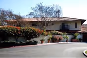 Single-story building with a terracotta roof, landscaped shrubs, and potted plants along a curved driveway.