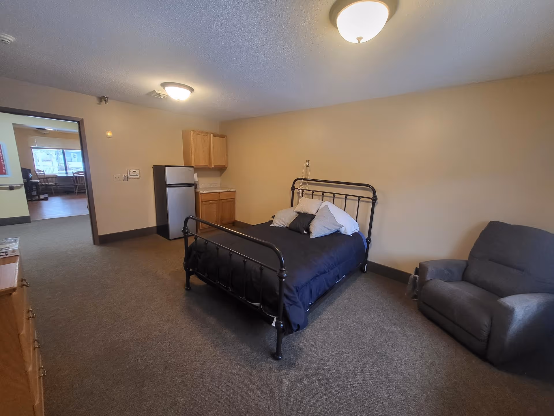 A simple bedroom in an assisted living facility featuring a black metal bed frame with pillows and a dark comforter, a gray armchair, a small kitchenette area with a mini refrigerator and wooden cabinets, and an open doorway leading to another room with tables and chairs.