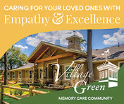 Exterior view of Village Green Memory Care Community Tomball building with a clear blue sky and some clouds. The building has a wooden roof structure and is surrounded by trees and greenery.