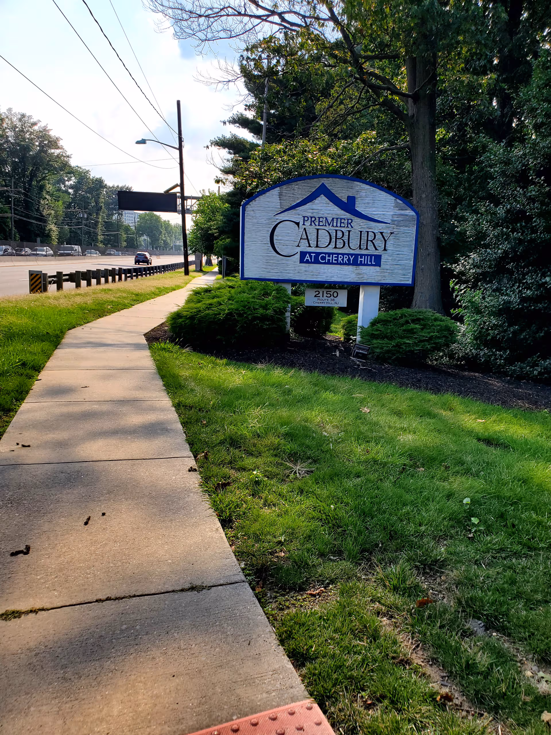 Sidewalk along a road with a Cadbury at Cherry Hill sign and trees.
