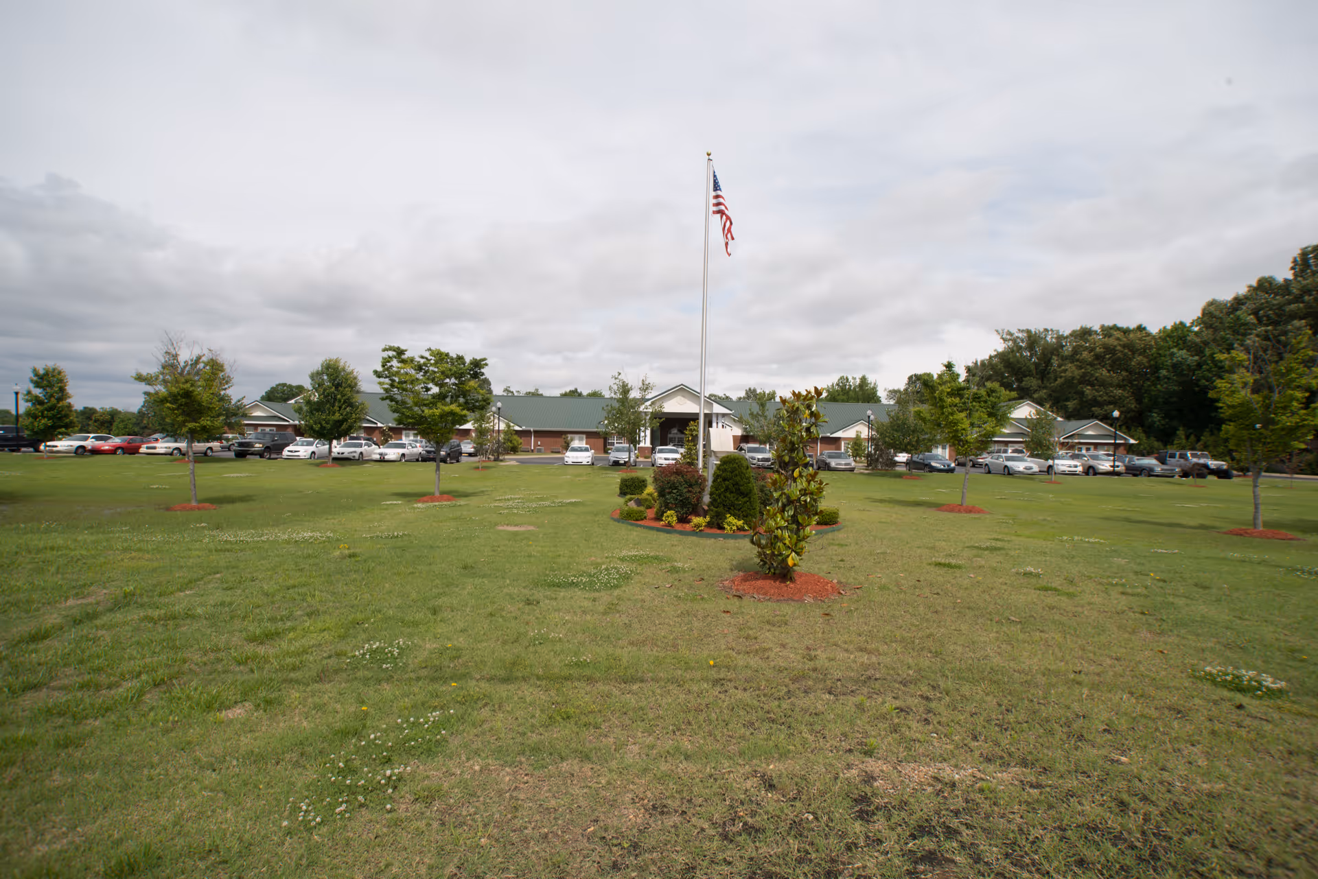 Wide grassy lawn with a central flagpole and landscaped beds in front of a one-story senior living facility building and parked cars under a cloudy sky.