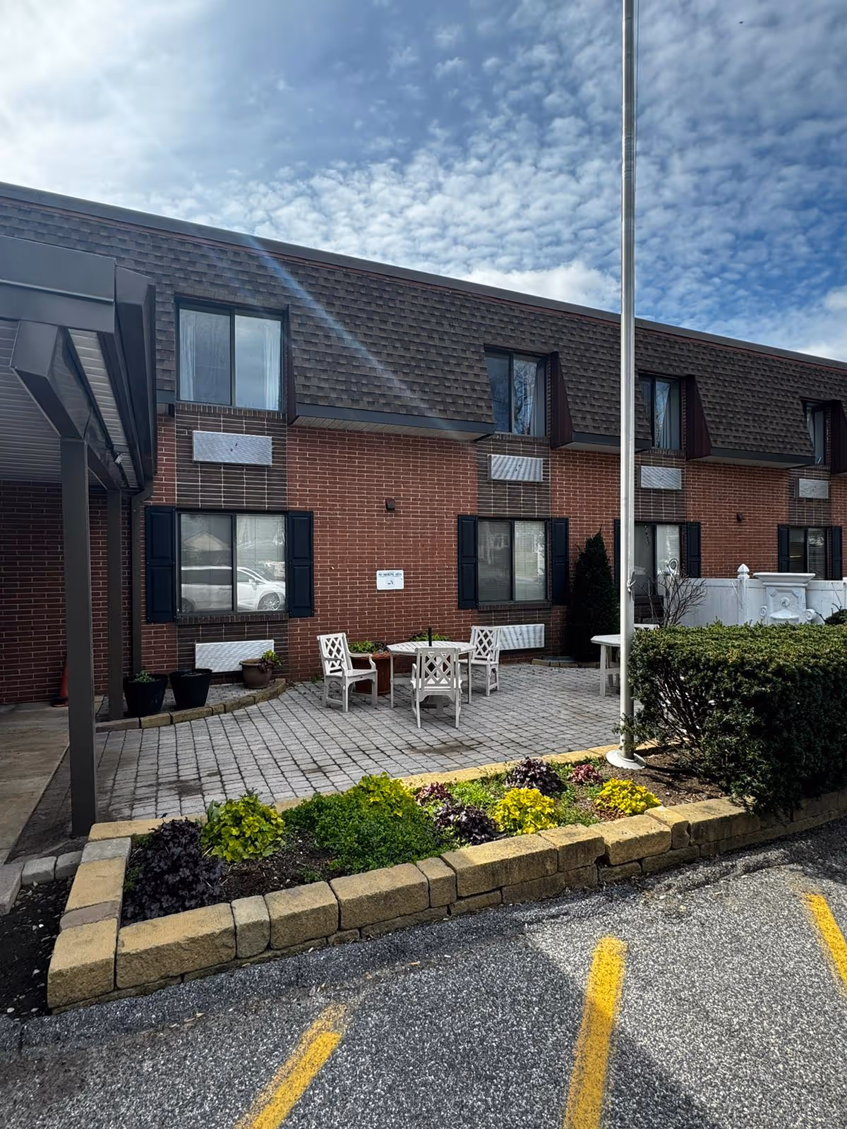 Brick two-story facility exterior with a paved patio area, white chairs and table, a flagpole, and a small flowerbed in front.