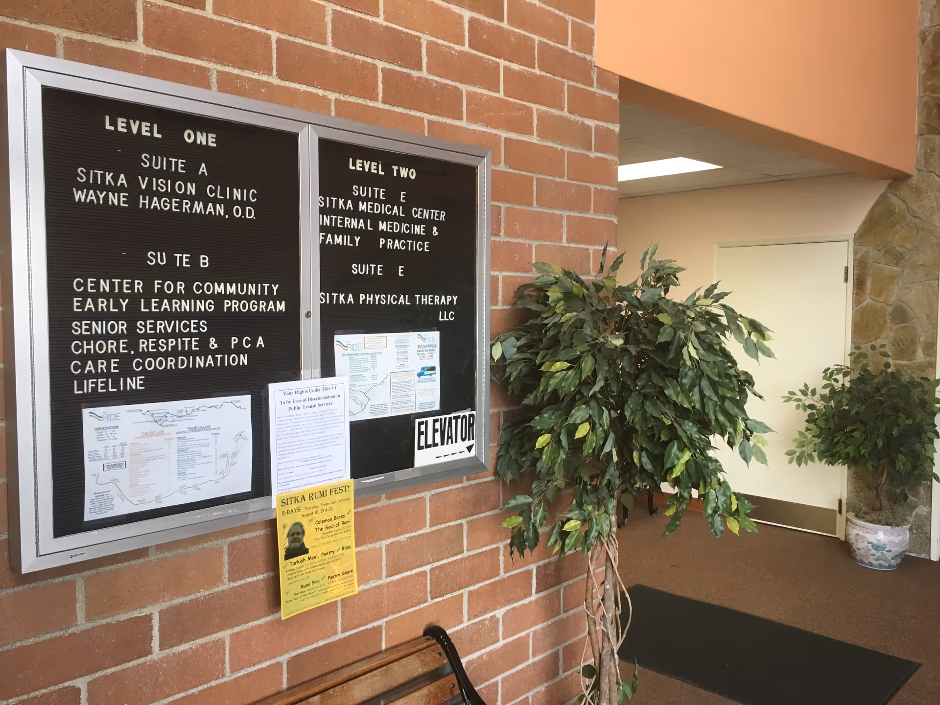 Brick-walled interior lobby with bulletin boards, elevator, doorway, and potted plants.