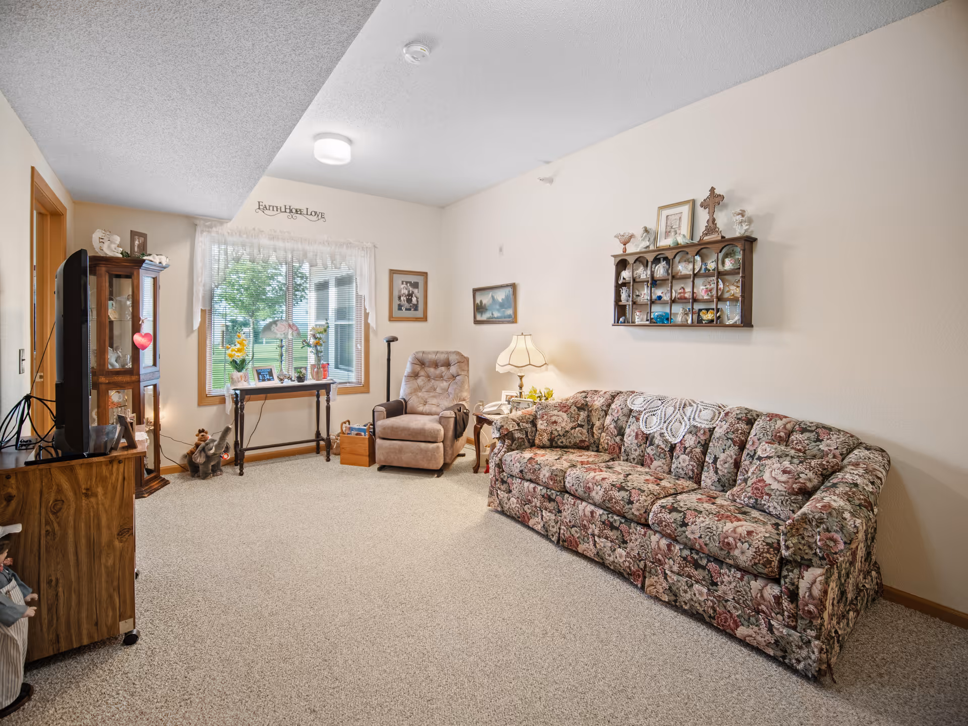 A cozy living room with a floral patterned sofa, a beige recliner chair, a wooden side table with a lamp, and a small table under a window with lace curtains. The walls are decorated with framed pictures and a wooden shelf displaying various decorative items. The room has beige carpet and soft lighting.