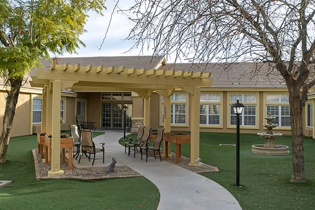Outdoor seating area with a wooden pergola, several chairs, and a concrete pathway leading through a grassy courtyard. The courtyard is surrounded by a building with multiple windows, a lamppost, a water fountain, and some trees.