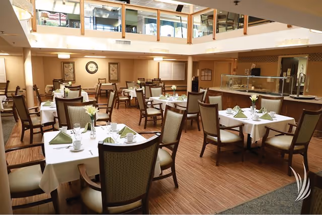 A spacious dining room with multiple tables covered in white tablecloths, each set with green napkins, cups, and small flower vases. The room has wooden flooring, cushioned chairs, and a serving area with a glass sneeze guard. There is a mezzanine level with glass railings above the dining area.
