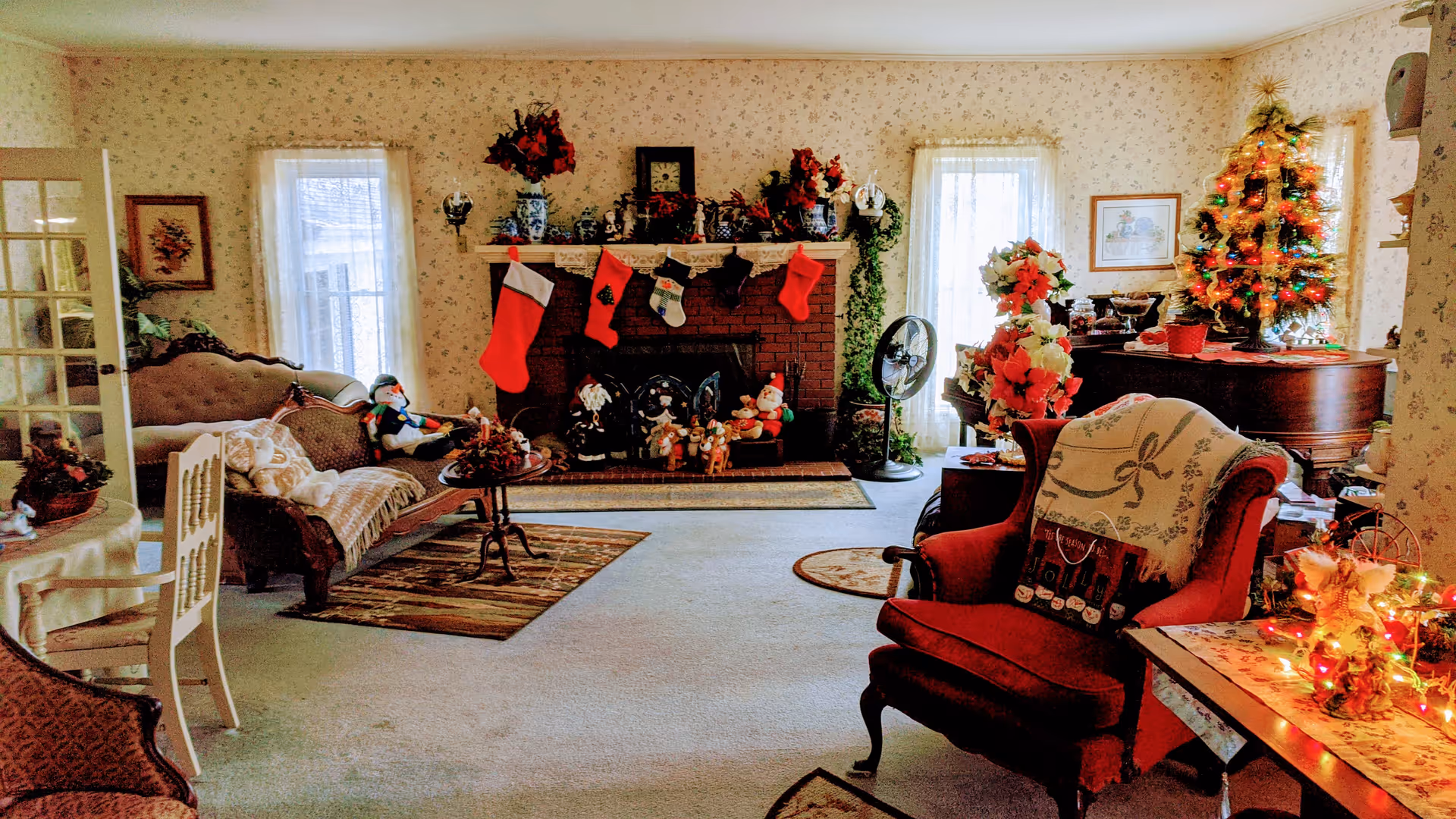 Cozy living room decorated for Christmas with a brick fireplace, stockings, seating, and a small lit tree.