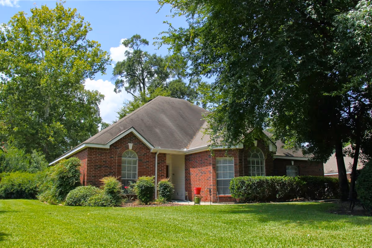 A single-story brick house with a sloped roof surrounded by green grass, bushes, and tall trees under a blue sky with some clouds.