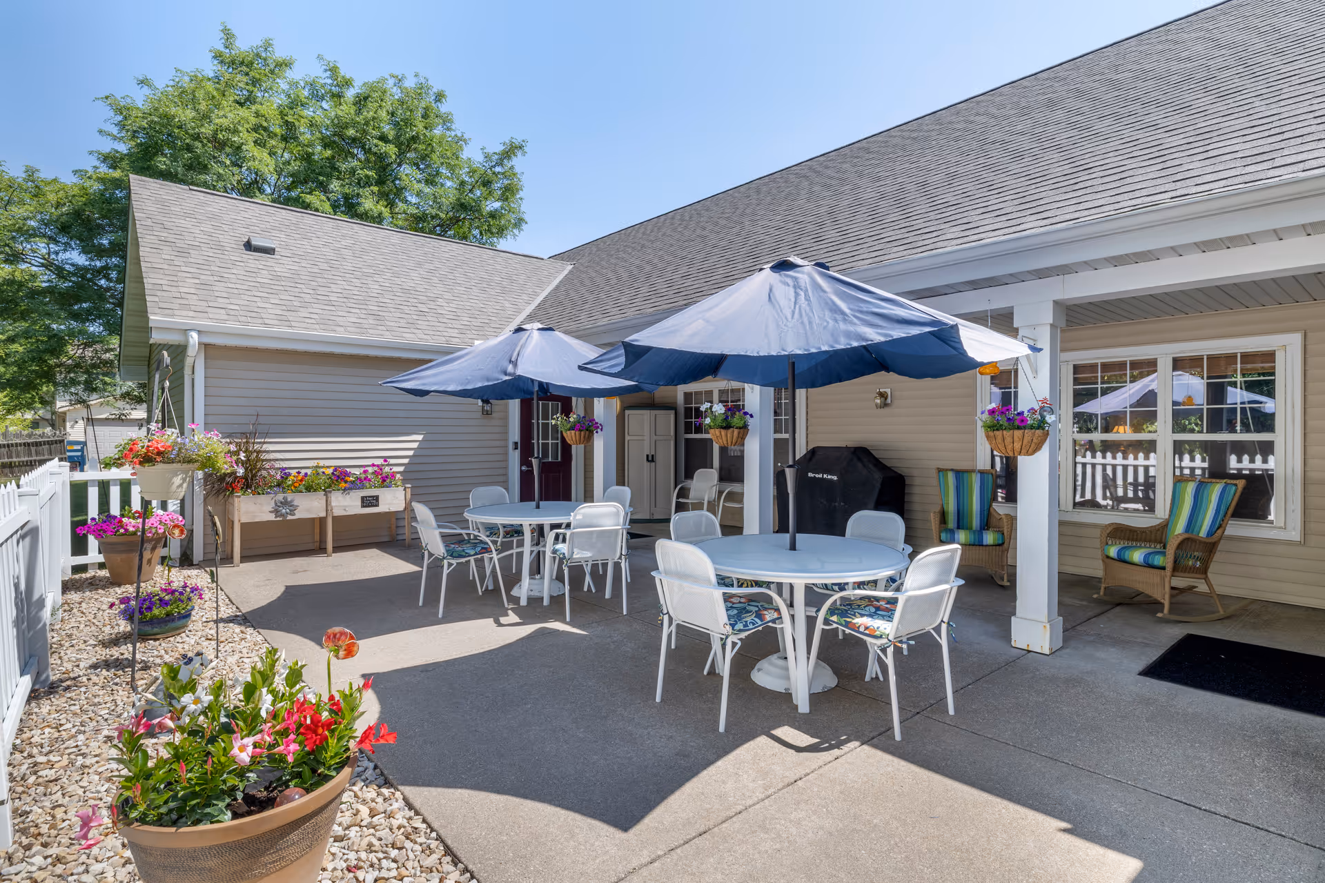 Outdoor sunlit patio with round tables, blue umbrellas, white chairs, potted flowers and lounge seating beside a beige building.