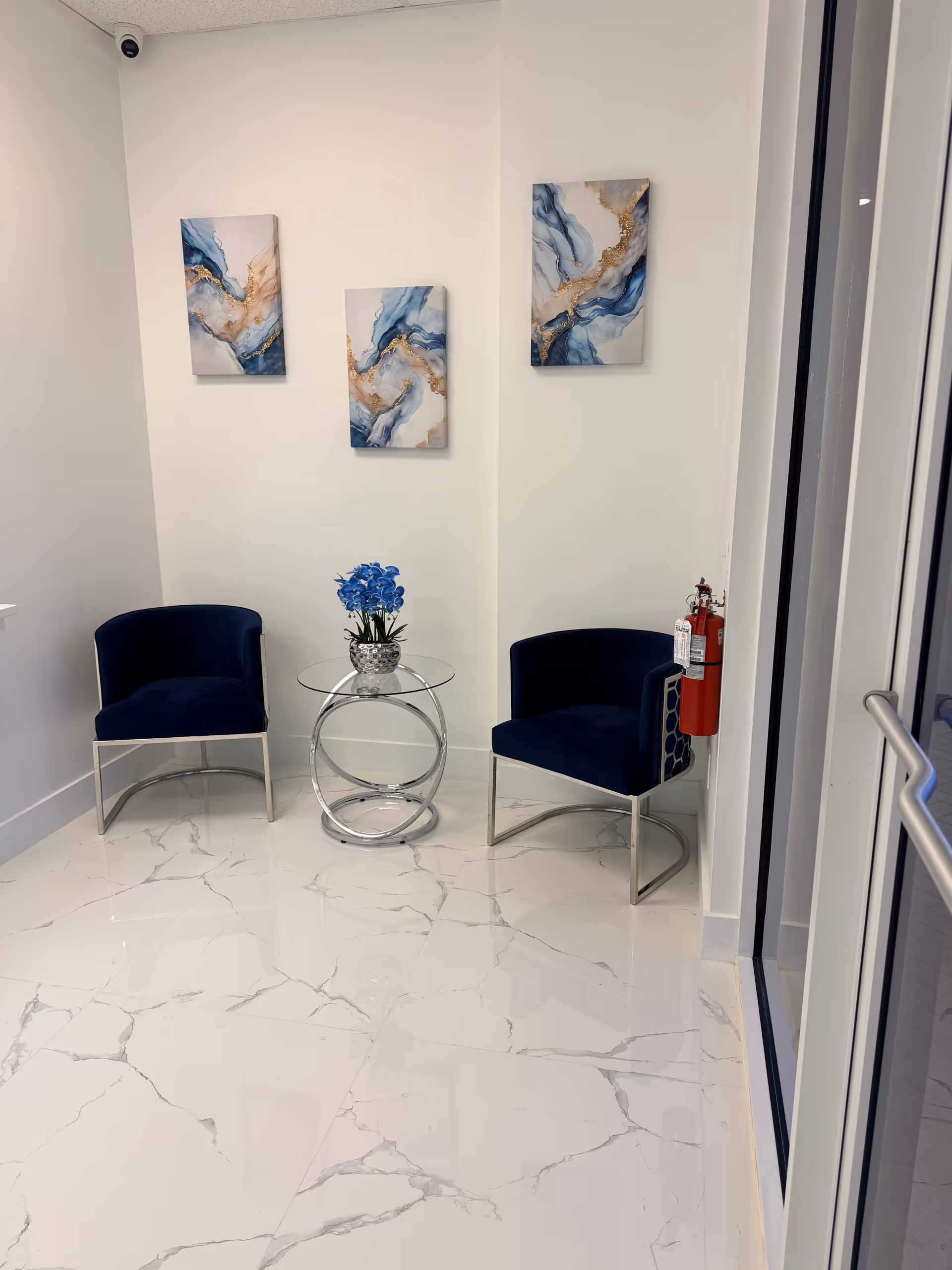 A modern waiting area with two blue chairs, a glass table, and wall art.