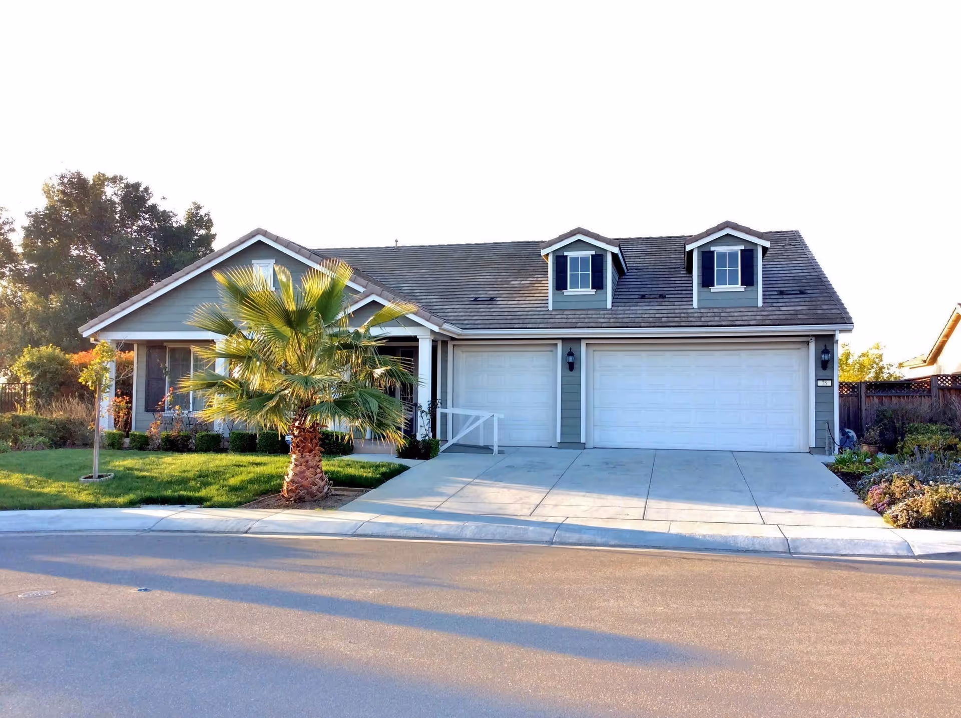 Exterior view of a single-story residential building with a gray roof and light green siding. The house features two dormer windows with black shutters, a double garage with white doors, a small front porch, and a palm tree in the front yard. The driveway and street are visible in the foreground.