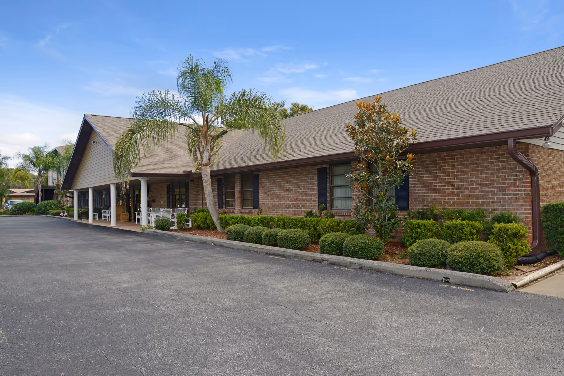 Brick senior living building with a covered entrance, palm trees, and manicured shrubs.