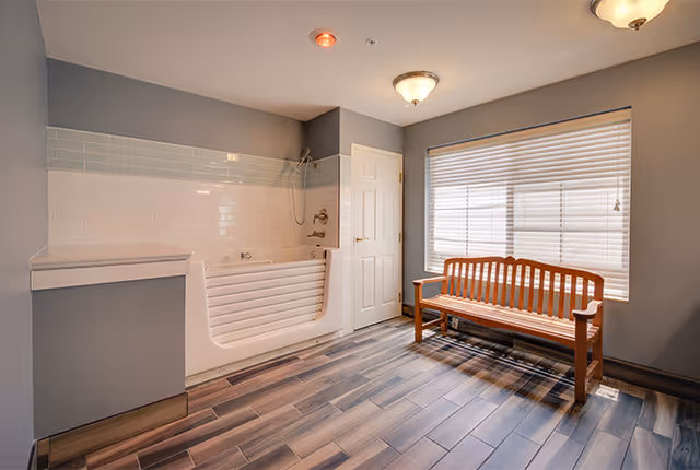 A spacious bathroom featuring a walk-in bathtub with a handheld showerhead, a wooden bench near a large window with blinds, and wood-look tile flooring. The walls are painted gray with white tile accents around the bathtub.