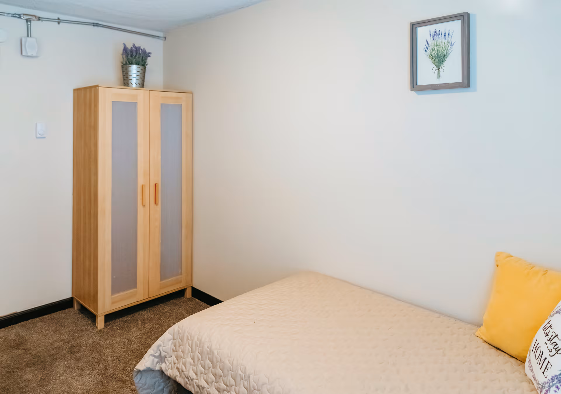 A simple bedroom with a single bed covered in a beige quilt, a yellow pillow, and a decorative pillow with text. There is a wooden wardrobe with frosted glass panels in the corner and a small potted plant on top. A framed picture of lavender hangs on the wall above the bed.
