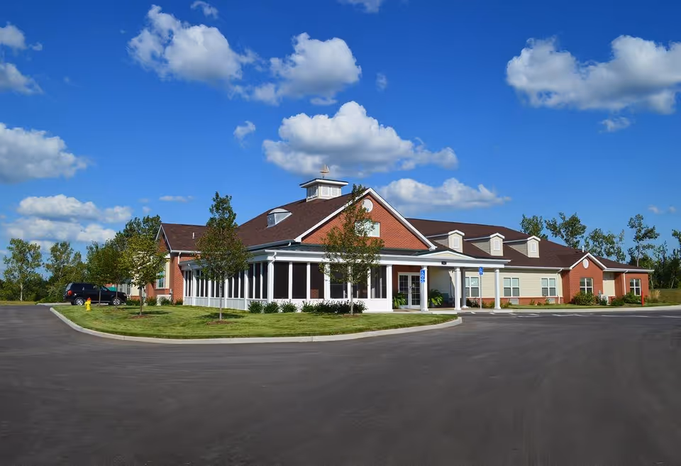Exterior view of a single-story brick and siding building under a blue sky with scattered clouds. The building has a covered entrance with white columns and a circular driveway with parking spaces. There are small trees and grass landscaping around the building.