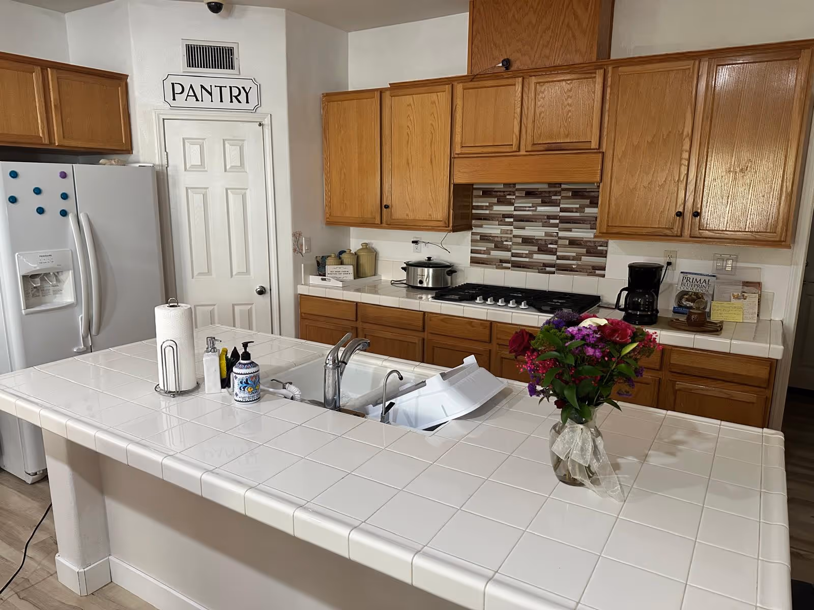 Bright kitchen with white tile island, wooden cabinets, stove, fridge, and flowers.