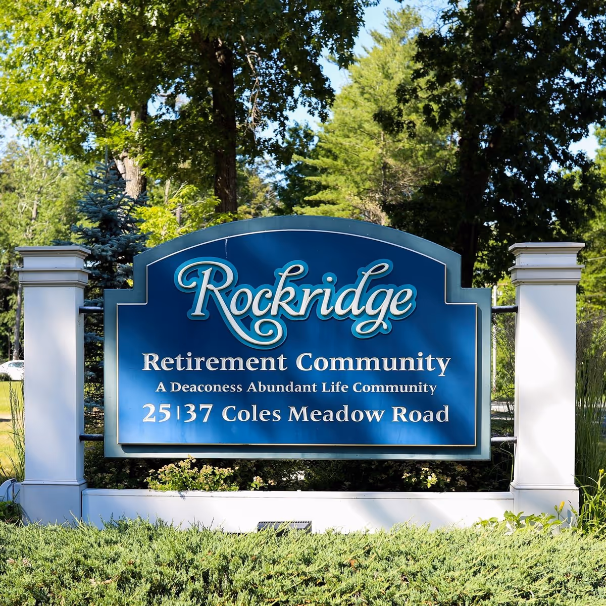 Blue Rockridge Retirement Community sign with white pillars and lush greenery in sun.