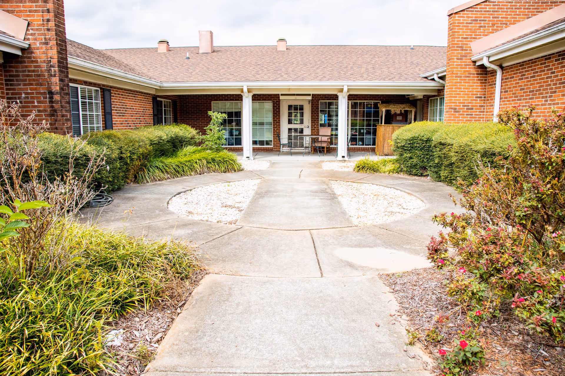 Concrete walkway leading through a landscaped courtyard to the covered entrance of a brick nursing facility with seating.