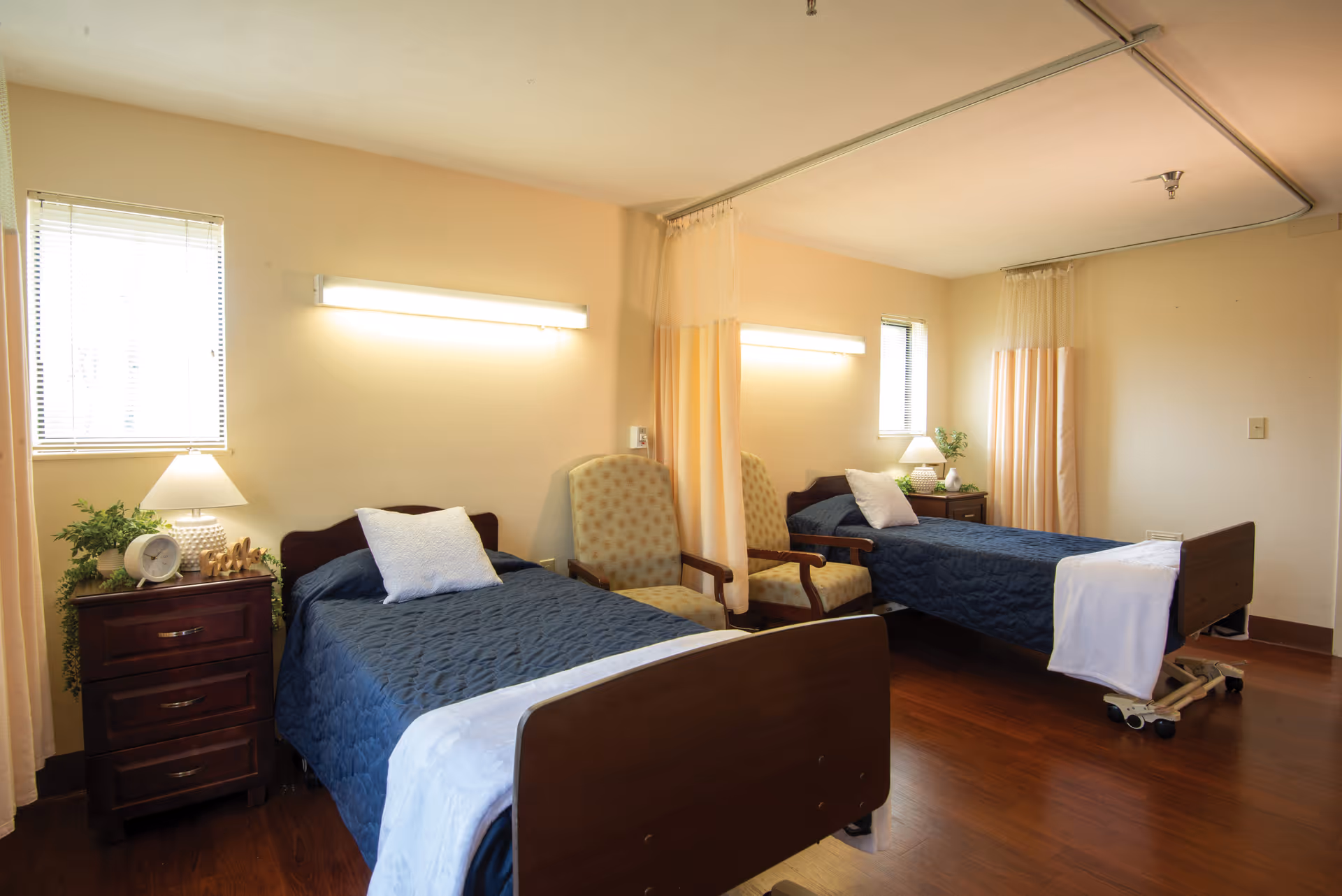 Shared senior living bedroom with two hospital-style beds, bedside tables, armchairs, lamps, and privacy curtains.