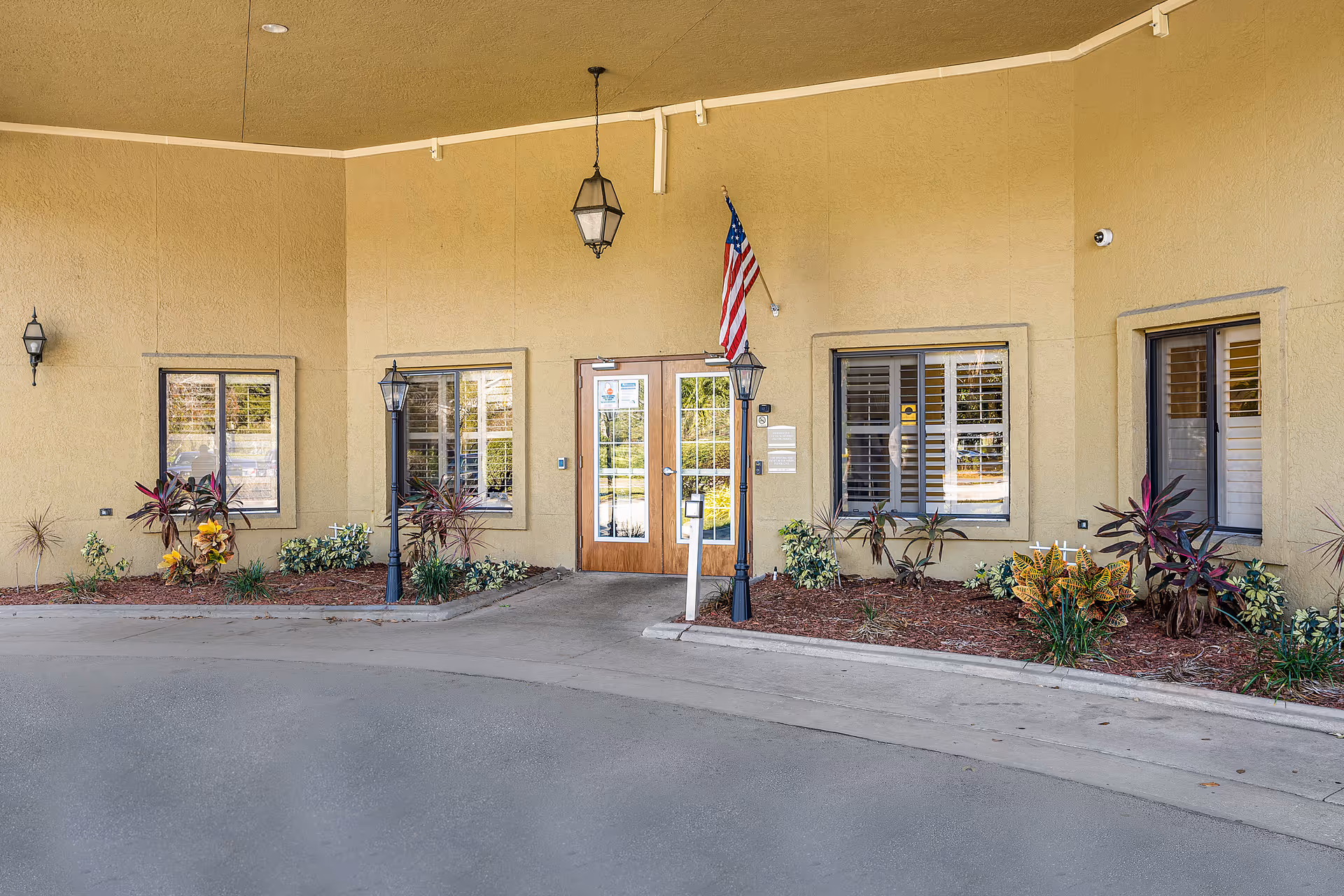 Covered front entrance of a building with double glass doors, an American flag, lantern-style lights, windows with shutters, and landscaped beds.