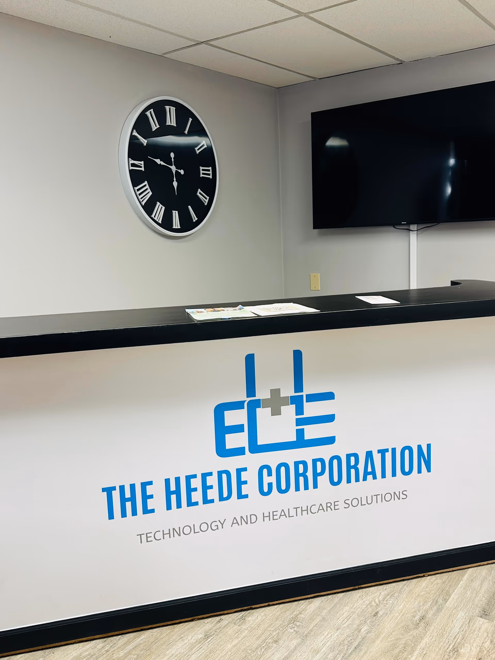 Reception desk with The Heede Corporation logo, clock on the wall, and a large TV.