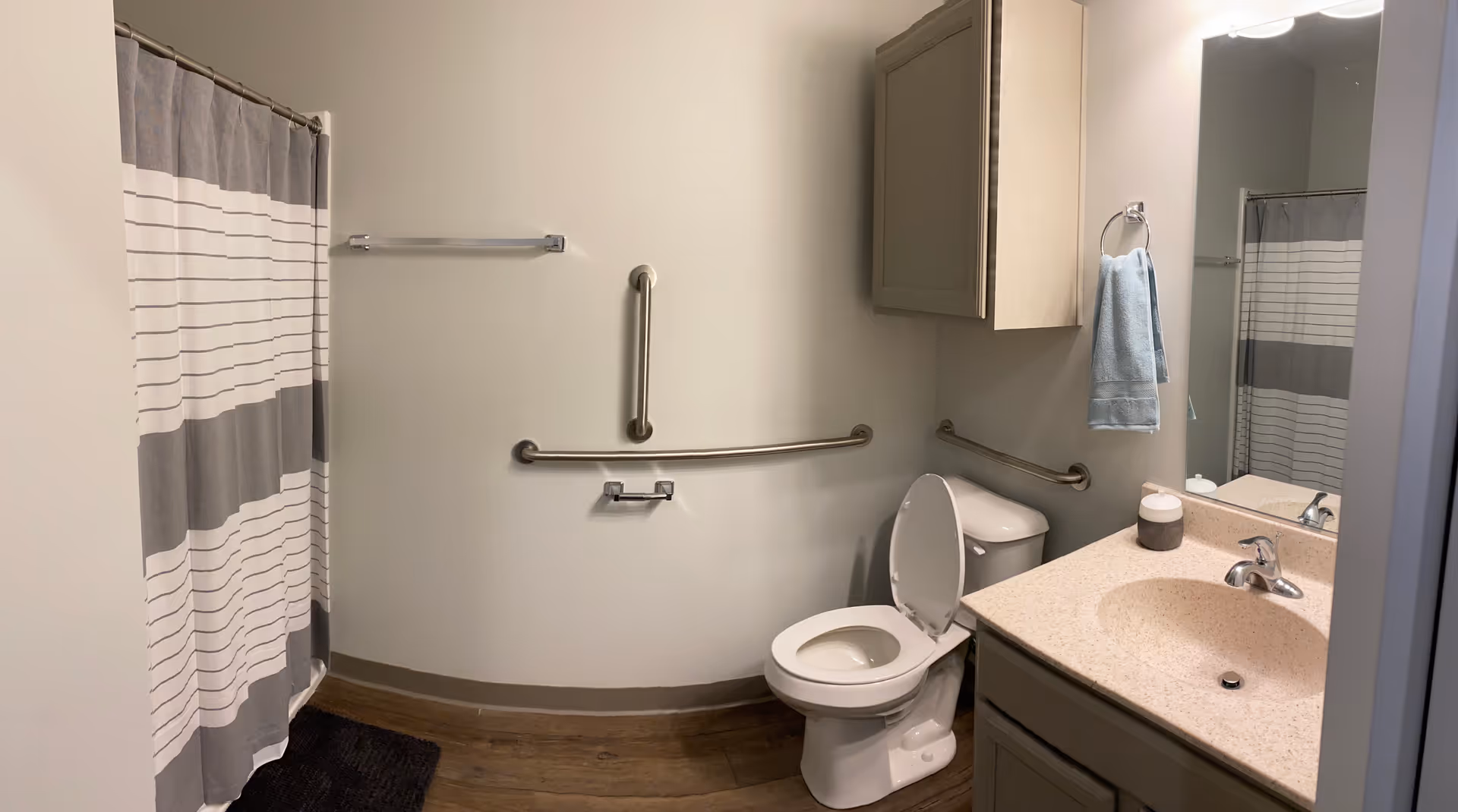 A clean and accessible bathroom with a toilet, grab bars on the walls, a sink with a countertop, a mirror, a towel hanging on a ring, a cabinet above the toilet, and a shower with a striped curtain.