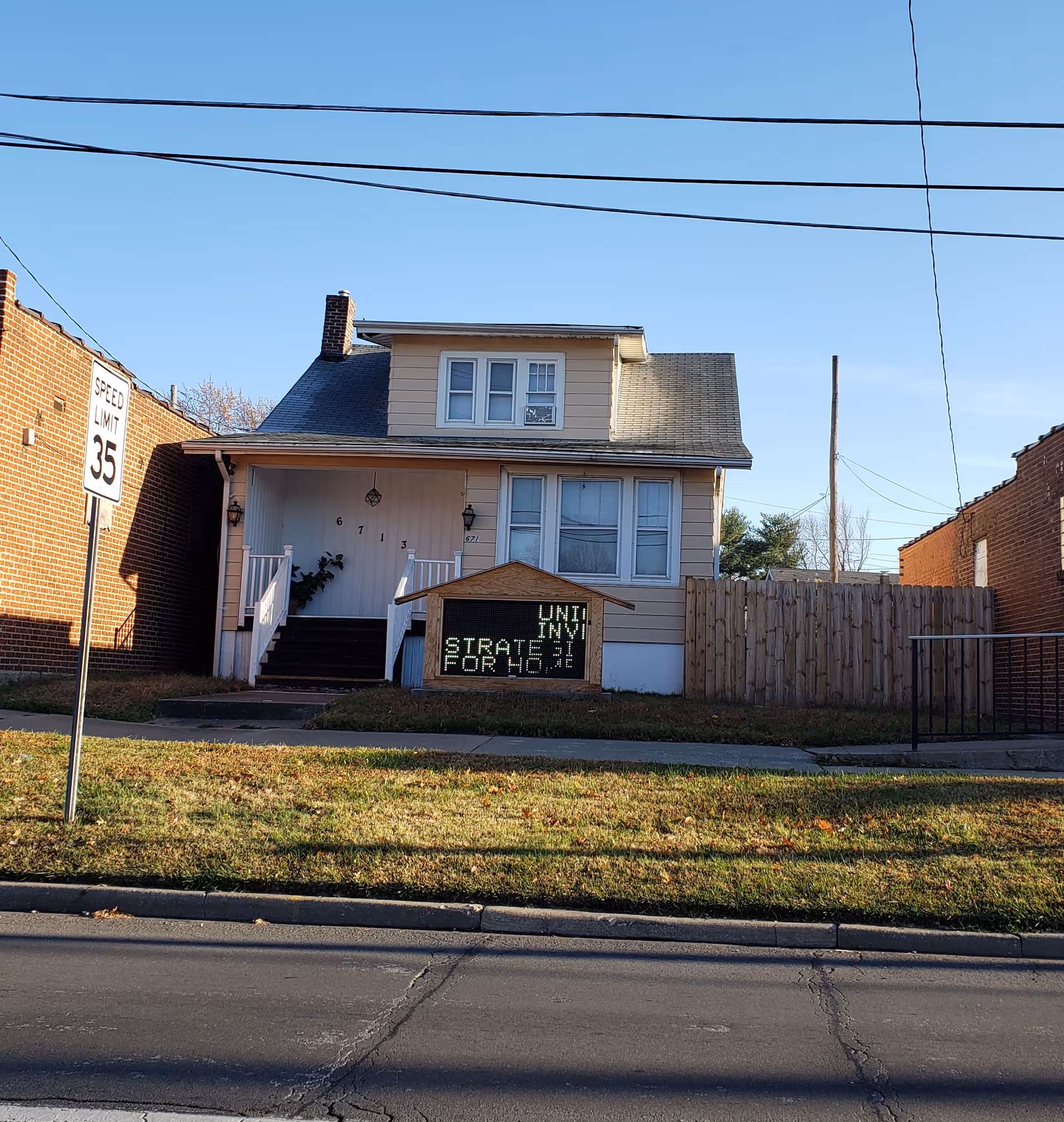 Two-story beige house with front porch, steps, and a signboard on the lawn.