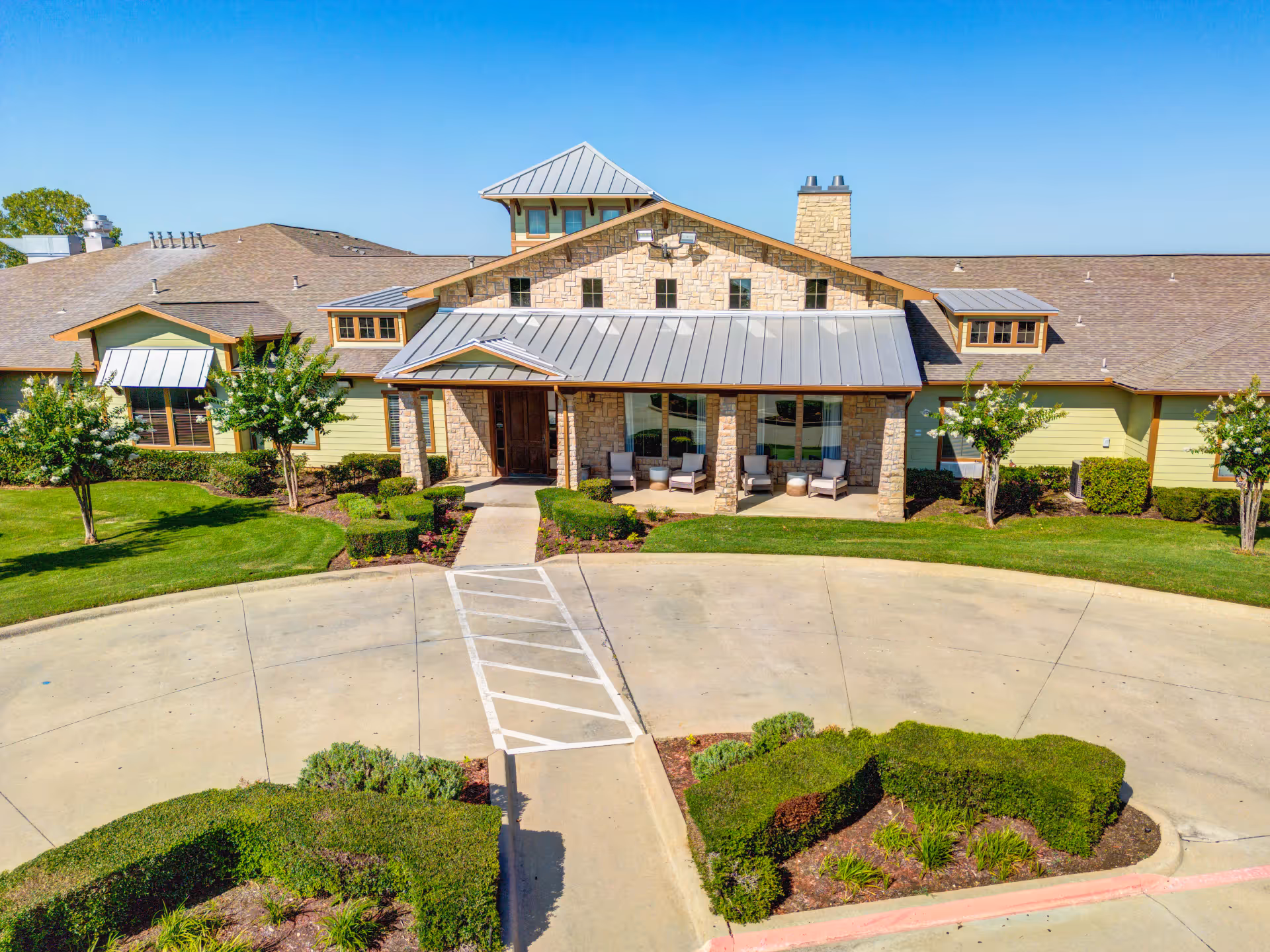 Front exterior view of a single-story assisted living facility building with a stone facade and a metal roof. The entrance has a covered porch with chairs and small tables. The surrounding area is landscaped with green grass, bushes, and small trees under a clear blue sky.