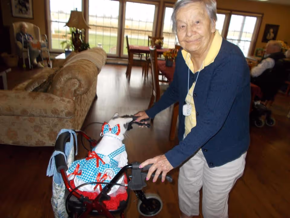 An elderly woman smiling and standing indoors, holding a walker with a small dog dressed in a blue and white checkered outfit with red bows sitting on it. The room has wooden floors, a beige patterned couch, and large windows in the background letting in natural light. Other elderly individuals are seated in the background.