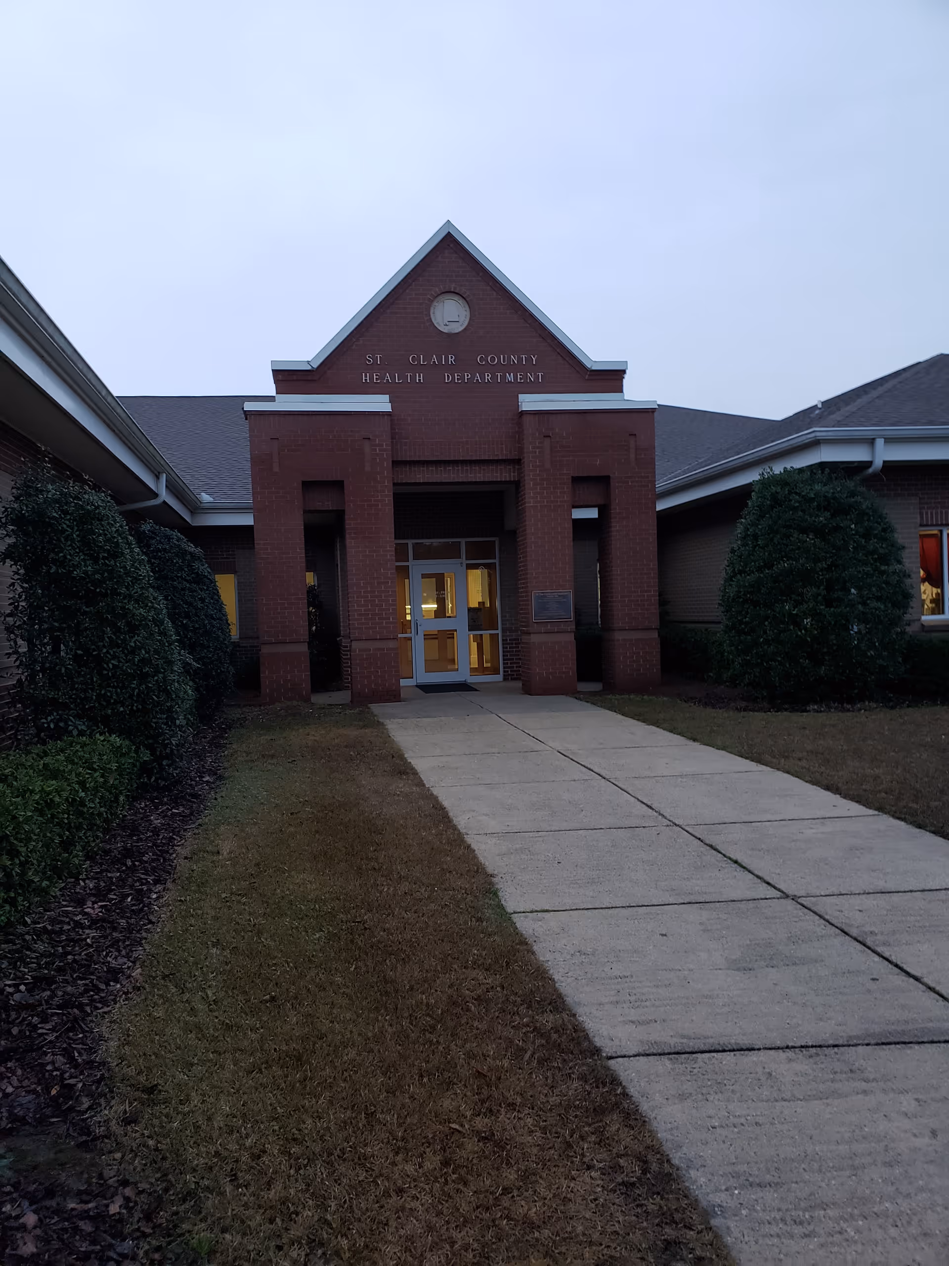 Front entrance of a brick St. Clair County Health Department building with a walkway and shrubs.