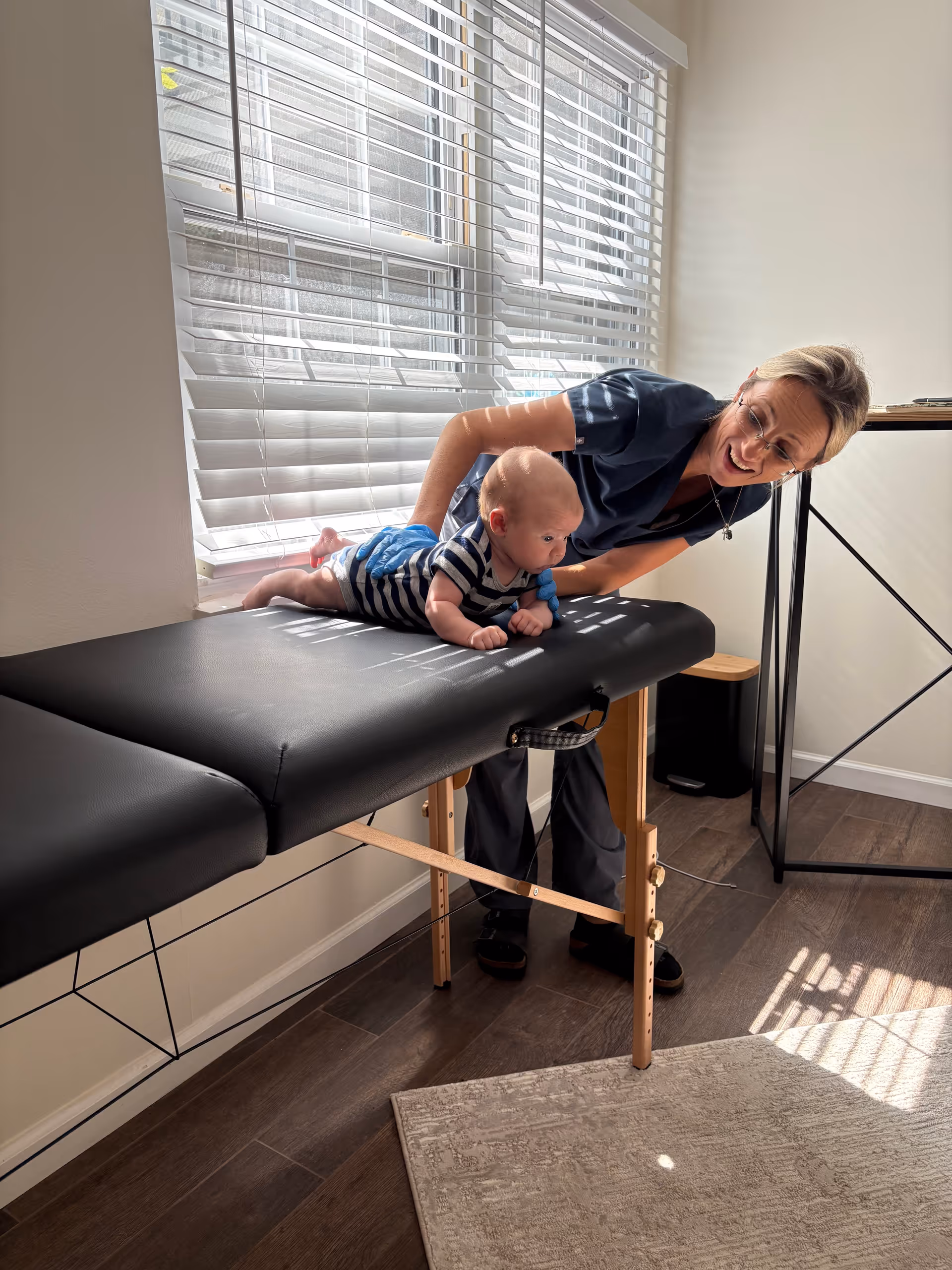Caregiver leans over a baby on a black examination table by a window.