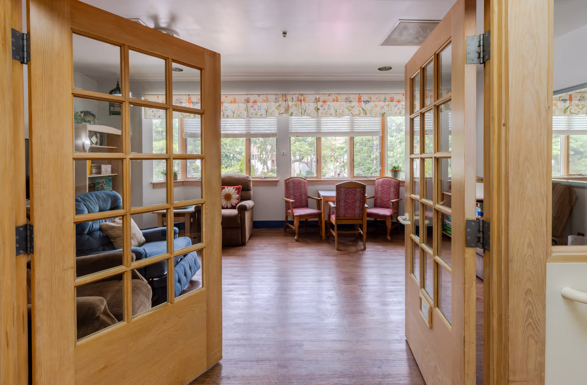 Open double wooden doors lead to a bright common living room with seating and windows.