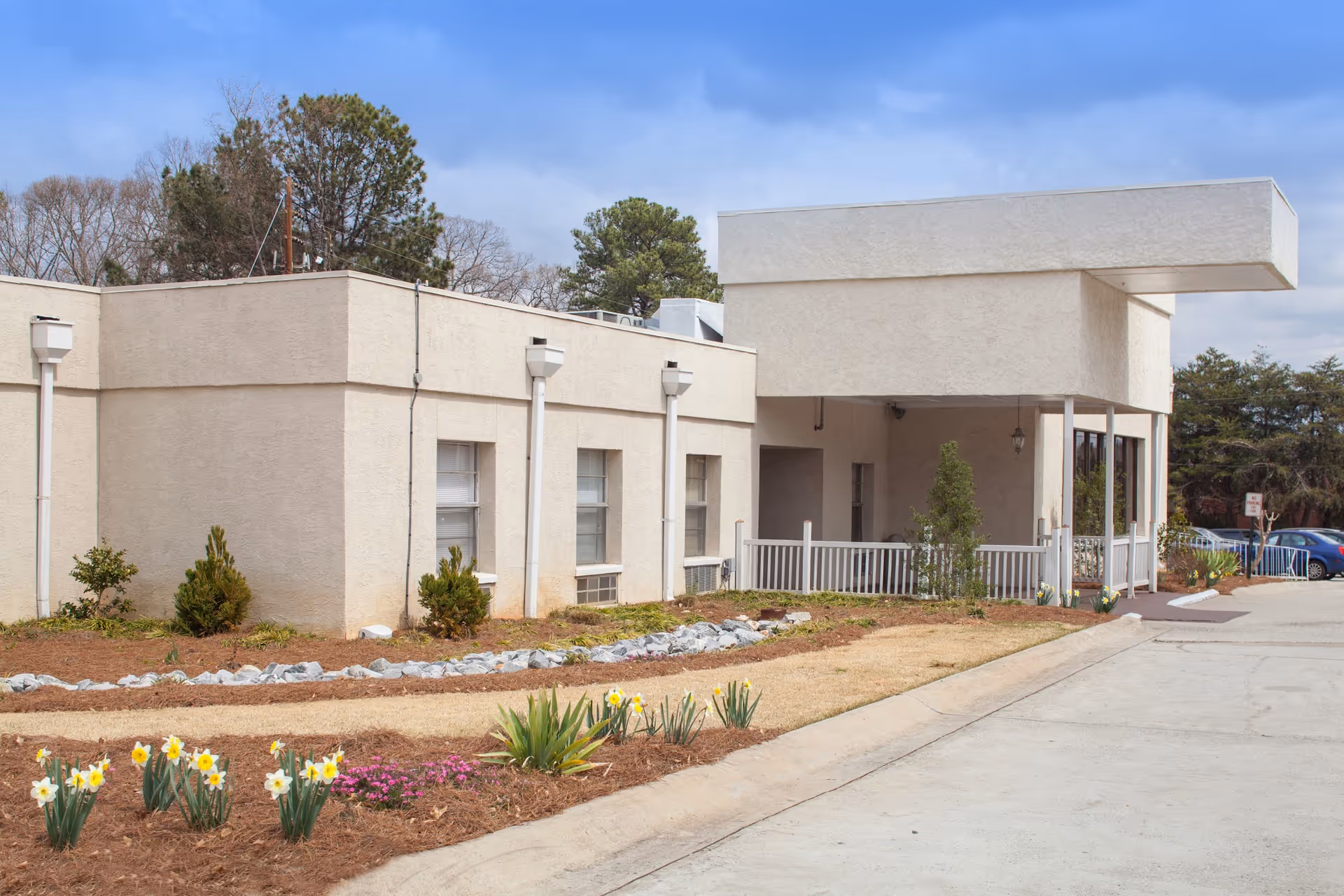 Exterior view of a single-story beige building with a covered entrance, small shrubs, and flower beds with daffodils and other plants along a curved pathway. Several trees are visible in the background under a partly cloudy sky.