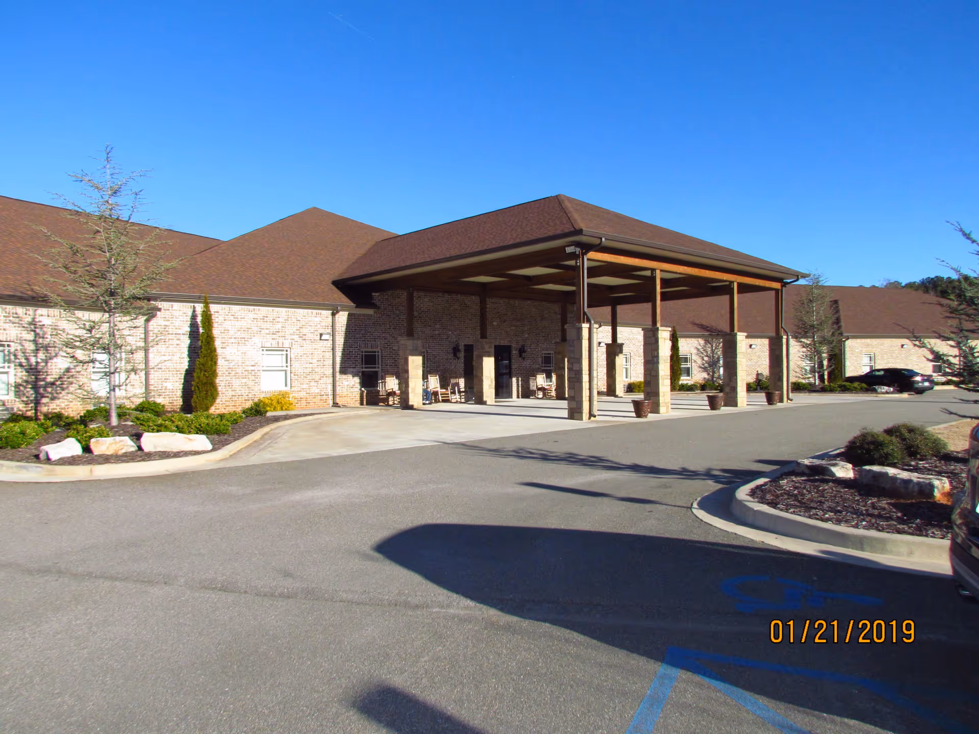 Front entrance of a single-story brick assisted living building with a covered drop-off porte-cochere, driveway and landscaped beds under a clear blue sky.
