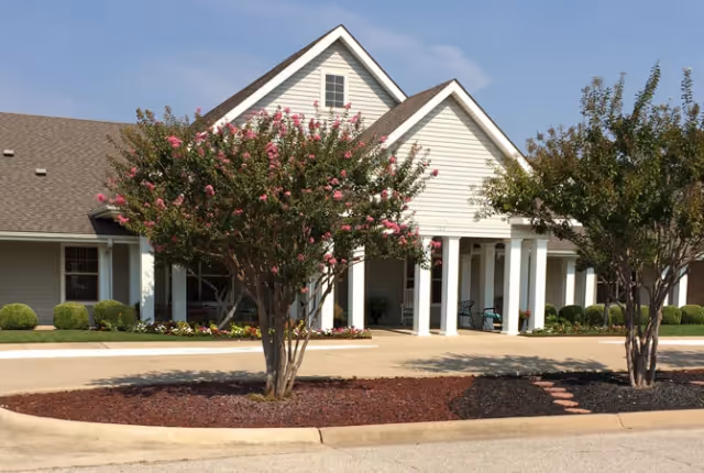 Front entrance of a light-colored residential building with white columns, a porch, and flowering trees in landscaped beds.
