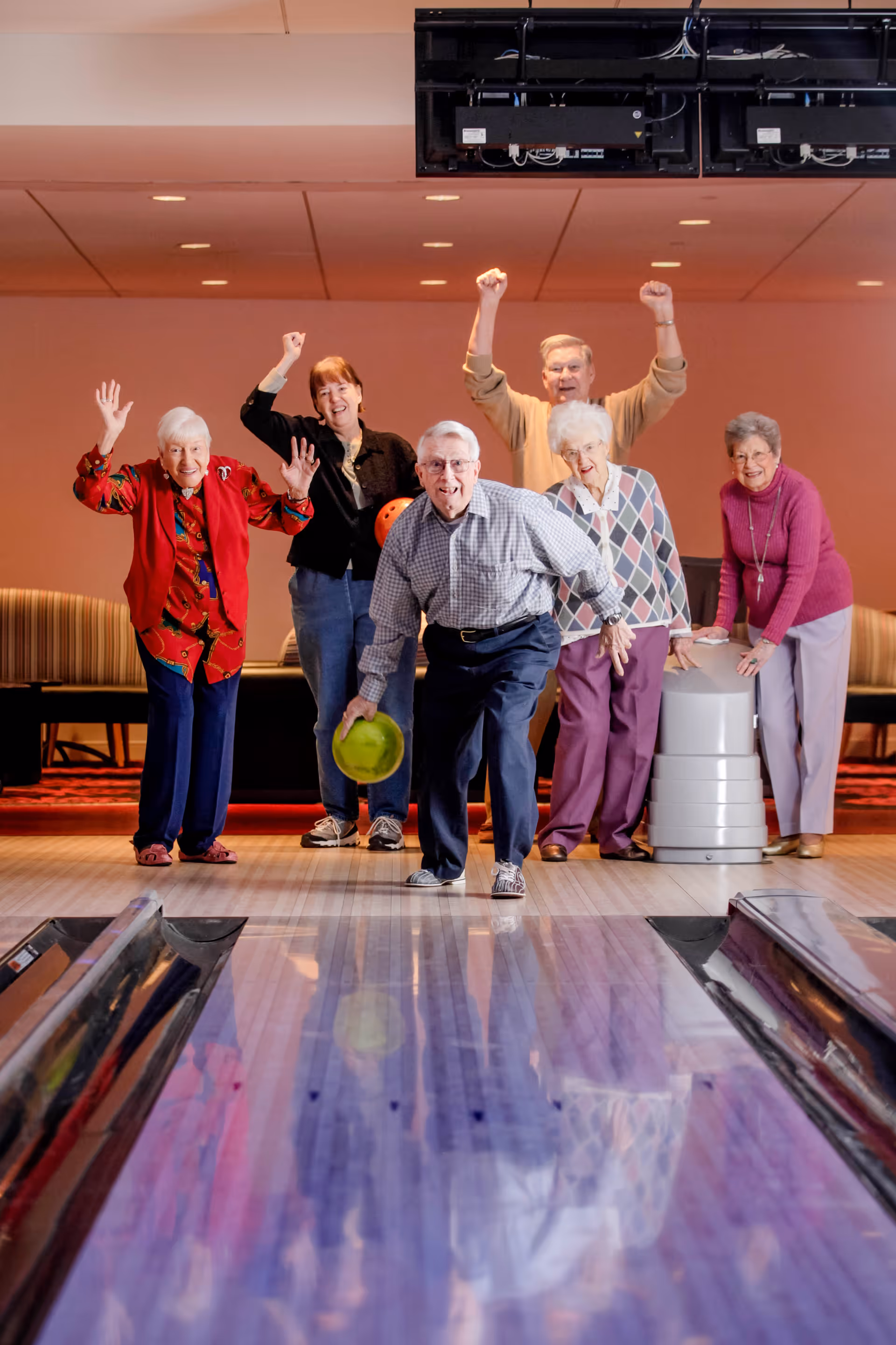 A group of six elderly people enjoying a game of bowling indoors. One man in the front is about to roll a green bowling ball down the lane while the others cheer and smile behind him.