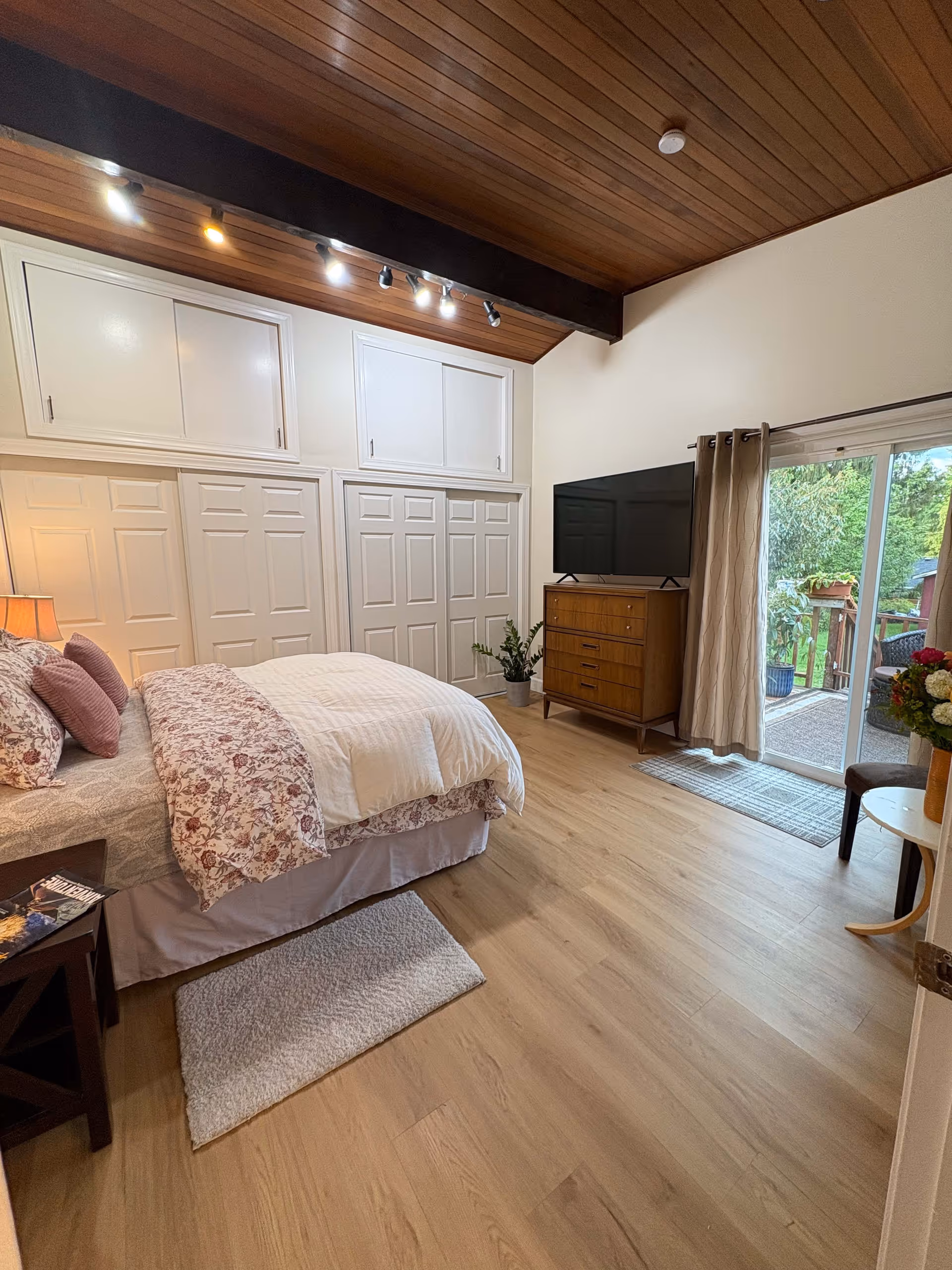 A cozy bedroom with a wooden ceiling and light wood flooring. The room features a bed with floral and white bedding, a bedside table with a lamp and a magazine, a wooden dresser with a flat-screen TV on top, and a sliding glass door leading to an outdoor area with greenery. There are multiple white closet doors along one wall and a small rug on the floor beside the bed.