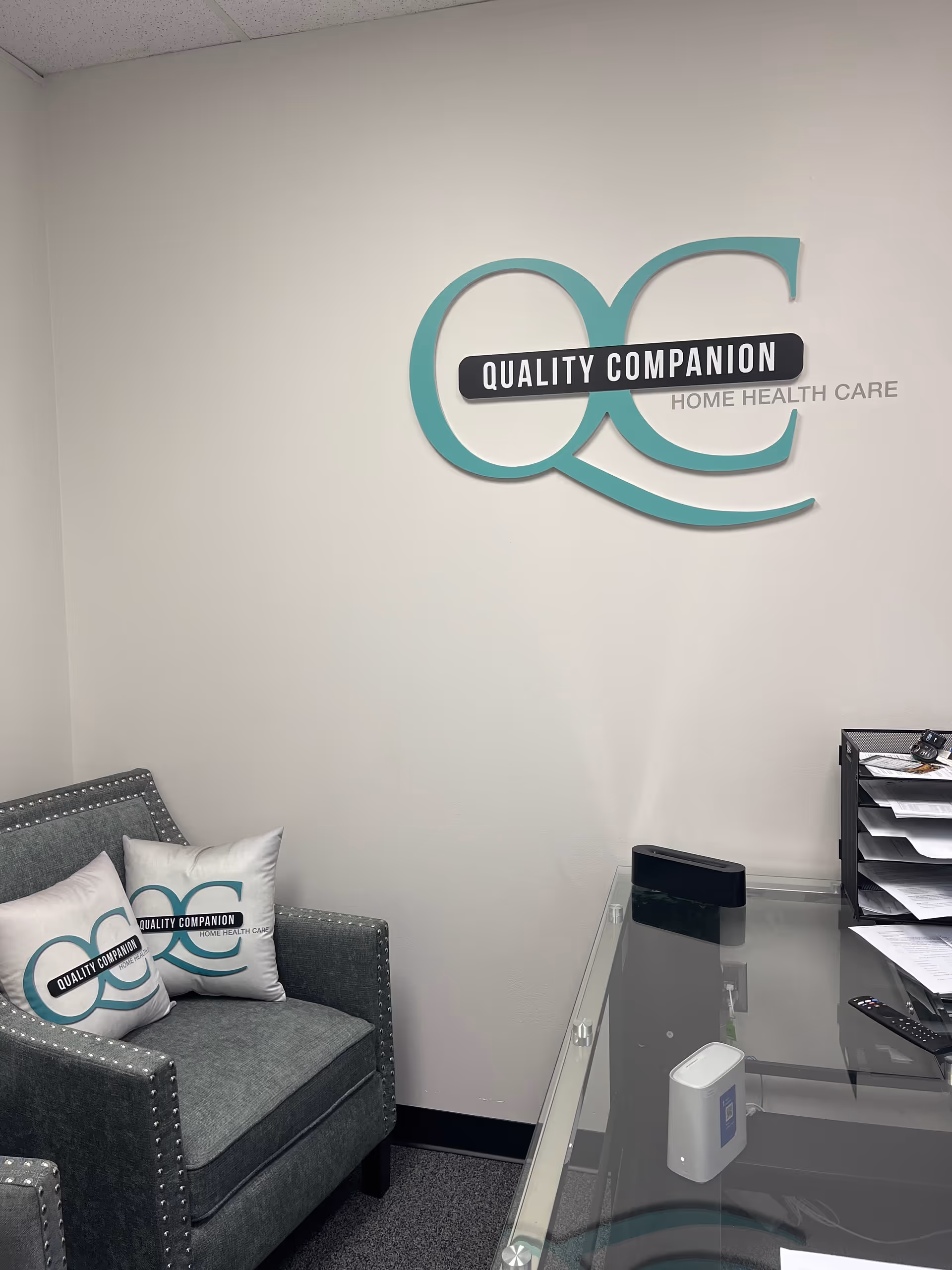 Office reception area with teal Quality Companion logo on wall, seating and glass desk.