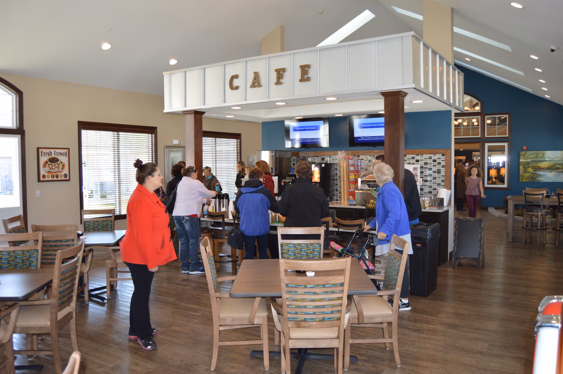 Interior view of a cafe area in an assisted living facility with several people gathered around the counter. The space features wooden tables and chairs, large windows with blinds, and a sign above the counter that reads 'CAFE'. The floor is wooden, and the walls are painted beige and blue. There are two TV screens mounted above the counter and a sign on the wall that says 'Fresh Brewed'.