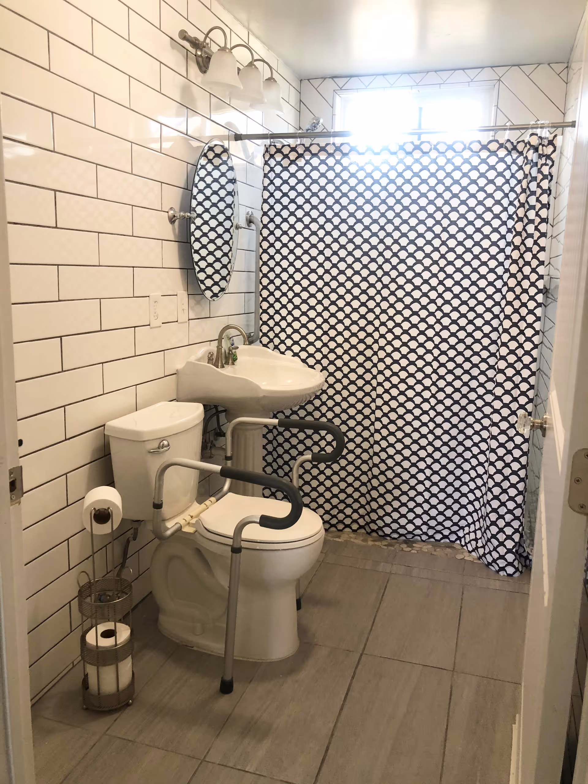 Small tiled bathroom with a toilet fitted with safety grab bars, a pedestal sink, and a patterned shower curtain.