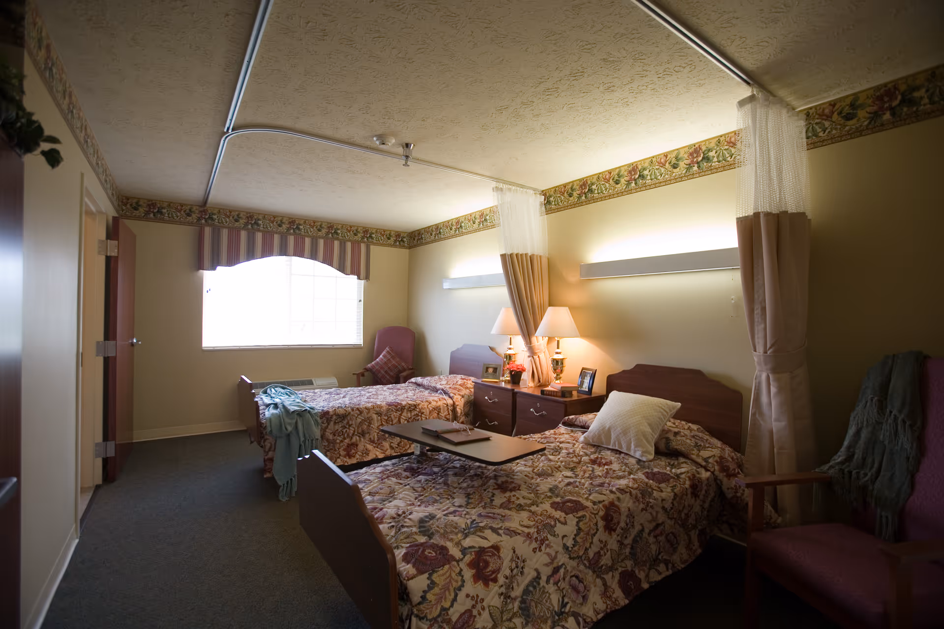 Two double beds with floral bedspreads, lamps, nightstands, and privacy curtains in a cozy bedroom.