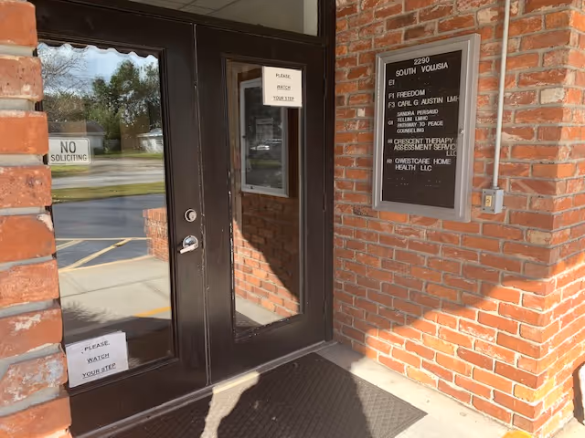 Brick building entrance with glass doors, signs, and a wall plaque, no people visible.