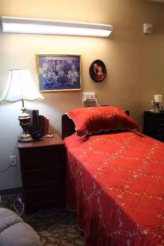 A cozy bedroom with a single bed covered in a red bedspread with floral patterns. Next to the bed is a wooden nightstand with a lamp, books, and a clock. Above the bed on the wall are two framed pictures, one rectangular family photo and one smaller oval portrait. The room has warm lighting and a carpeted floor.