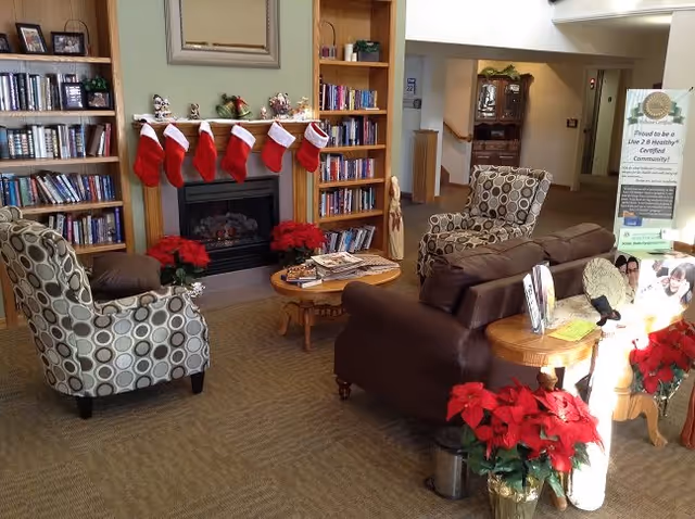 A cozy living room area with a fireplace decorated with six red Christmas stockings and poinsettia plants. There are two patterned armchairs and a brown leather sofa arranged around a wooden coffee table with magazines. Bookshelves flank the fireplace, and there is a standing sign in the background near a hallway.