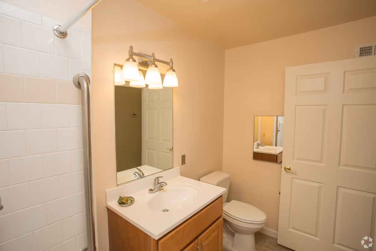 Small bathroom with vanity, sink, mirror, toilet, and shower in a senior living facility.